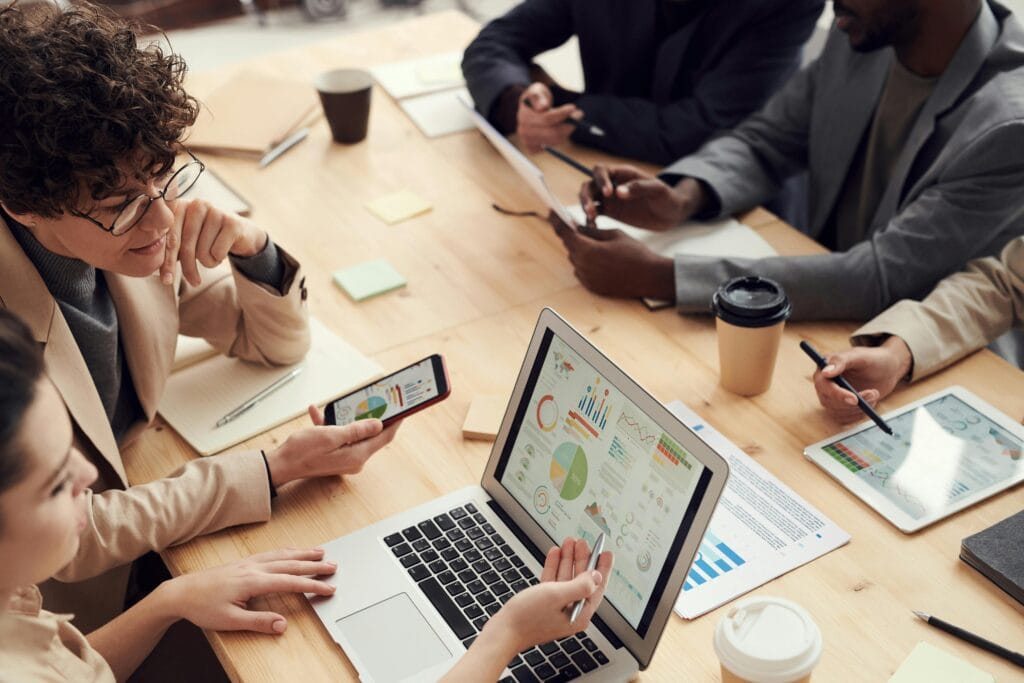 Grupo de pessoas em uma reunião de trabalho, concentradas em analisar gráficos e dados apresentados em dispositivos eletrônicos, como um laptop, smartphone e tablet. A mesa está repleta de documentos, blocos de notas e xícaras de café, indicando um ambiente de colaboração e discussão