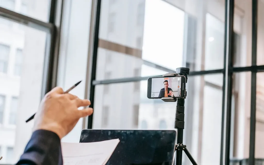 Fotografia de uma pessoa em um ambiente de escritório, participando de uma videoconferência ou gravação usando um smartphone montado em um tripé. A câmera do smartphone está voltada para a pessoa, que aparece na tela do dispositivo enquanto fala ou apresenta algo. A mão de outra pessoa, segurando uma caneta e um pedaço de papel, também aparece em primeiro plano, sugerindo que a pessoa está envolvida na conferência, talvez como espectadora ou participante.