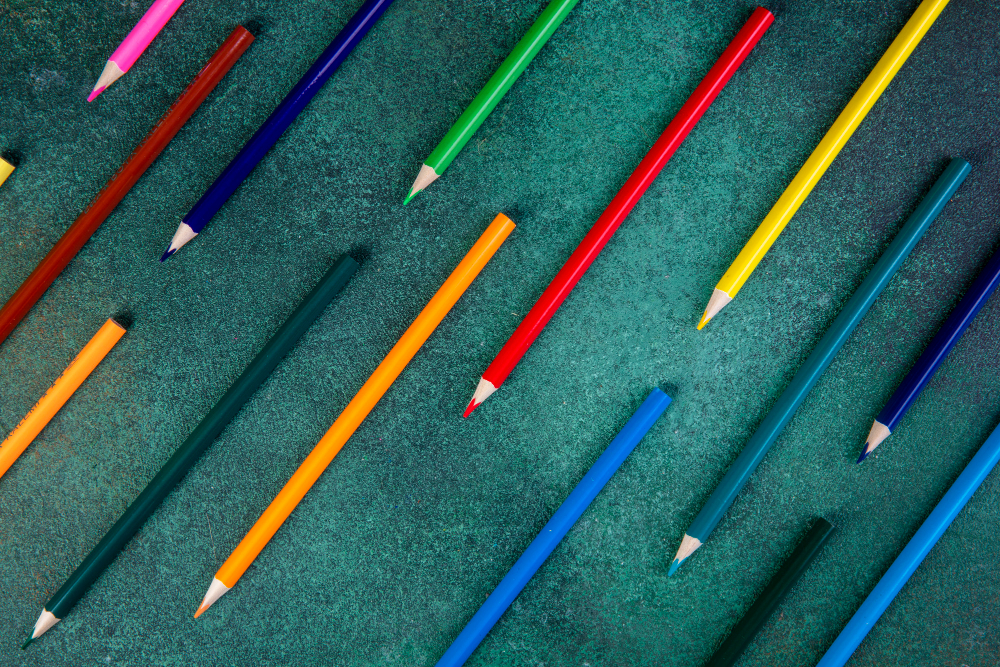 Diversos lápis de cor dispostos diagonalmente sobre um fundo verde texturizado, criando um padrão colorido e dinâmico