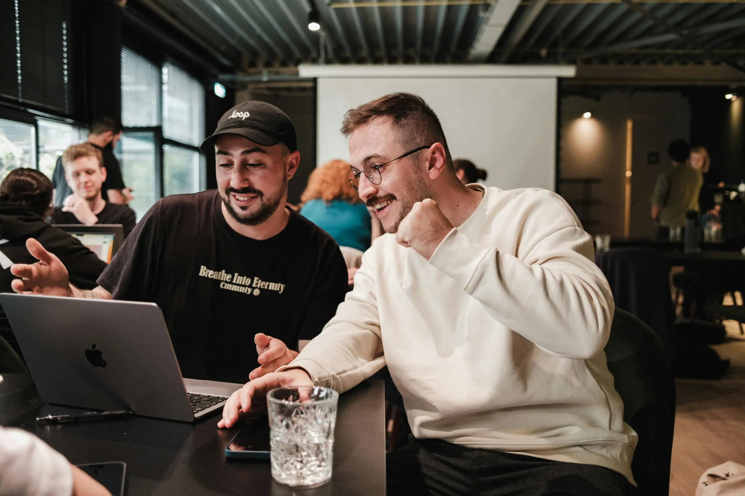 Duas pessoas estão sentadas lado a lado em uma mesa de trabalho, sorrindo enquanto interagem com um notebook. Uma delas usa boné e camiseta preta, e a outra veste um moletom branco e óculos. Ao fundo, outras pessoas também estão reunidas em um ambiente de coworking moderno e iluminado.